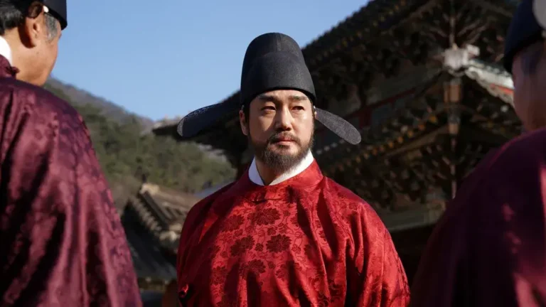 야외 궁궐 마당에서 붉은 관복과 검은 사모를 갖춰 입은 한명회를 연기하는 유지태 배우가 주변 인물들 사이에 서서 단호한 표정으로 앞을 바라보는 한국 사극 영화의 한 장면.