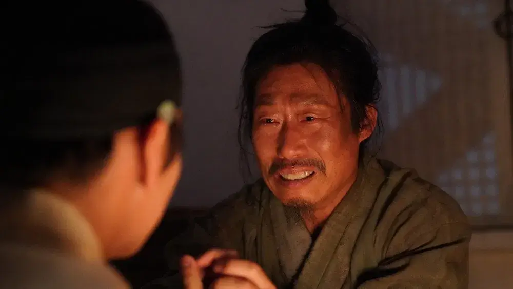 불빛이 비추는 실내에서 엄흥도가 맞은편에 앉은 어린 왕을 바라보며 긴장과 미소가 뒤섞인 표정을 짓는 영화 왕과사는남자의 한 장면.