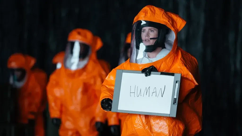 주황색 보호복을 입은 여성 과학자가 ‘HUMAN’이라고 적힌 보드를 들고 서 있고, 뒤로 같은 복장의 인물들이 보이는 SF 영화 컨택트의 상징적인 장면.