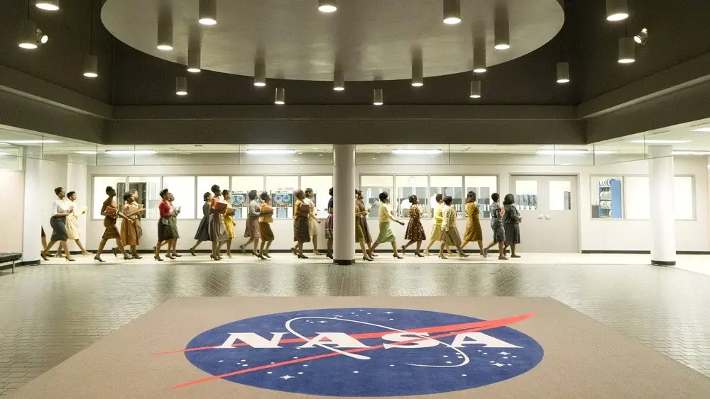 NASA 로고가 있는 넓은 실내 공간에서 여러 여성이 줄지어 이동하는 영화 _히든 피겨스_의 장면.