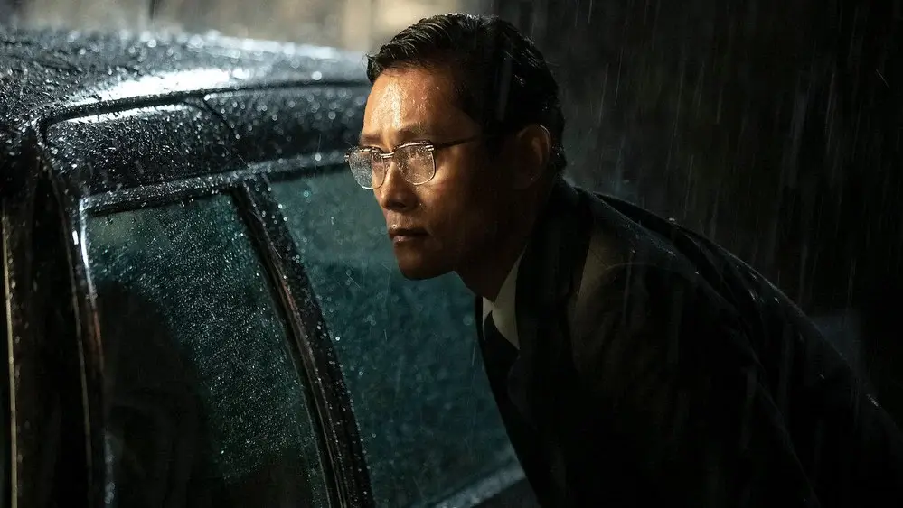비 내리는 밤, 검은 차 옆에 몸을 낮춘 안경 쓴 남성이 긴장한 표정으로 앞을 살피는 영화 _남산의 부장들_의 장면.