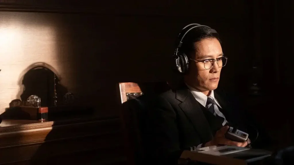어두운 실내에서 헤드폰을 쓴 안경 낀 남성이 녹음기를 손에 쥔 채 굳은 표정으로 앉아 있는 영화 남산의 부장들의 장면.