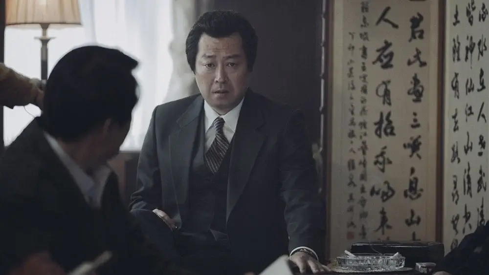1987 영화 스틸 이미지로, 박처원역을 연기하는 김윤석배우가 실내 회의 자리에서 당혹스럽고 무거운 표정으로 주변 인물들을 바라보는 장면