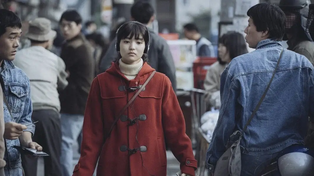1987 영화 스틸 이미지로, 연희역을 연기하는 김태리배우가 붉은 코트를 입고 혼잡한 거리 한가운데를 무표정하게 걸어가는 장면