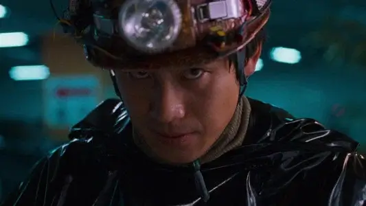 영화 지구를 지켜라 속 광부 헬멧과 검은 비닐 우비를 착용한 남성이 정면을 응시하는 장면으로, 집요하고 불안한 캐릭터의 에너지를 강조한 한국 영화 스틸 이미지