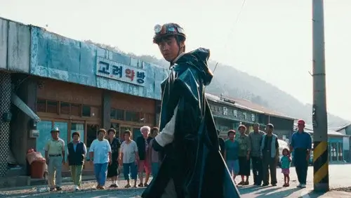 영화 지구를 지켜라 속 검은 우비와 헬멧을 쓴 남성이 시골 마을 한가운데 서 있고 주민들이 뒤에서 지켜보는 장면으로, 기묘한 영웅 서사와 블랙코미디 감성을 담은 스틸 이미지
