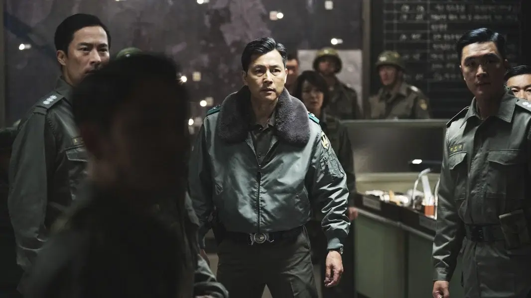 서울의 봄 영화 스틸 이미지로, 작전실 안에서 군 점퍼 차림 인물이 주변 장교들과 함께 긴급 상황을 마주하며 긴장감이 고조된 순간