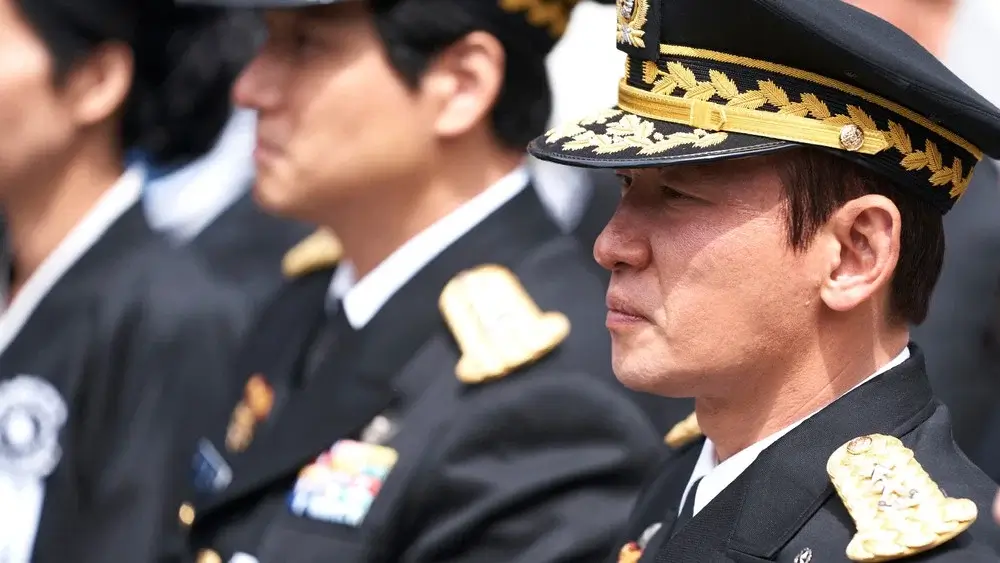 서울의 봄 영화 스틸 이미지로, 제복을 입은 고위 장성이 야외 행사장에서 엄숙한 표정으로 서 있는 군 권력 상징 장면