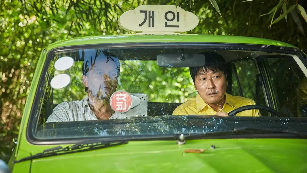 택시운전사 영화 스틸 이미지로, 김만섭역을 연기하는 송강호배우와 피터역을 연기하는 토마스 크레취만배우가 녹색 택시 안에 나란히 앉아 긴장된 표정으로 앞을 바라보는 장면