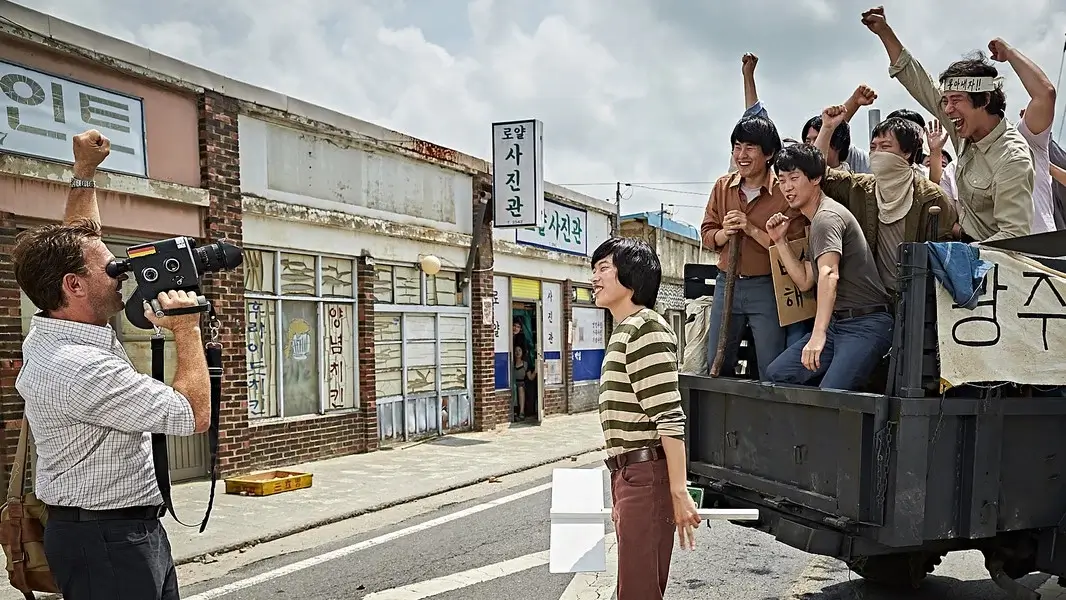택시운전사 영화 스틸 이미지로, 피터역을 연기하는 토마스 크레취만배우가 카메라를 들고 시민들 앞에서 촬영하고 김만섭역을 연기하는 송강호배우와 시민들이 함께 환호하는 장면