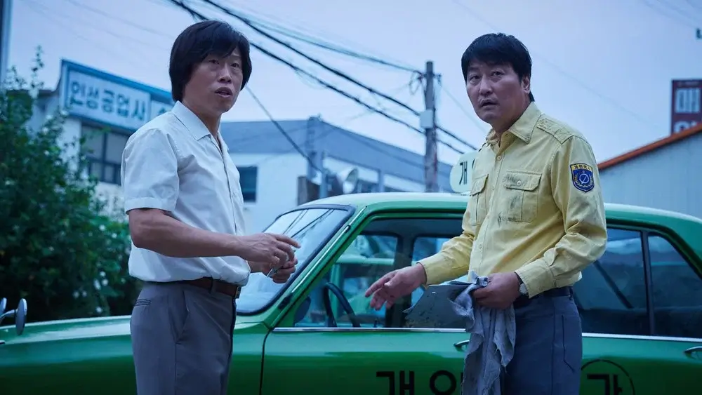 택시운전사 영화 스틸 이미지로, 김만섭역을 연기하는 송강호배우와 구재식역을 연기하는 류준열배우가 녹색 택시 앞에서 놀란 표정으로 상황을 지켜보는 장면