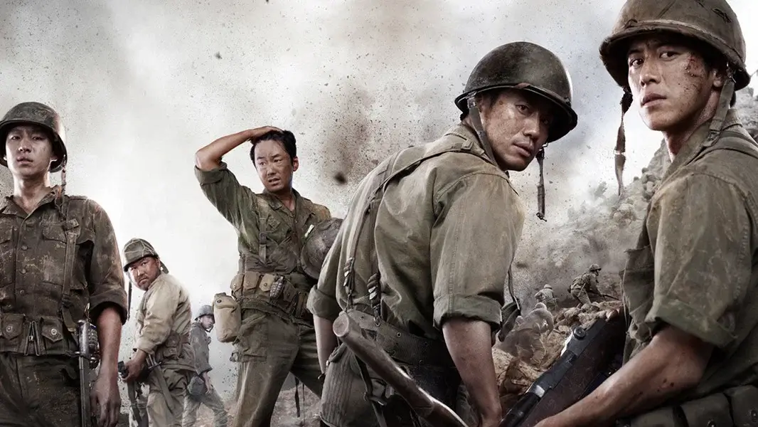고지전 영화 스틸 이미지로, 흙먼지와 폭발 흔적이 가득한 전장에서 여러 병사들이 뒤를 돌아보며 혼란과 생존 본능이 교차하는 전쟁 한복판의 분위기를 담은 장면