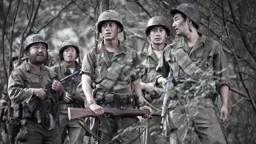 고지전 영화 스틸 이미지로, 숲속을 이동하던 병사들이 총을 든 채 주변을 살피며 적의 기습 가능성에 대비하는 긴박한 수색 장면