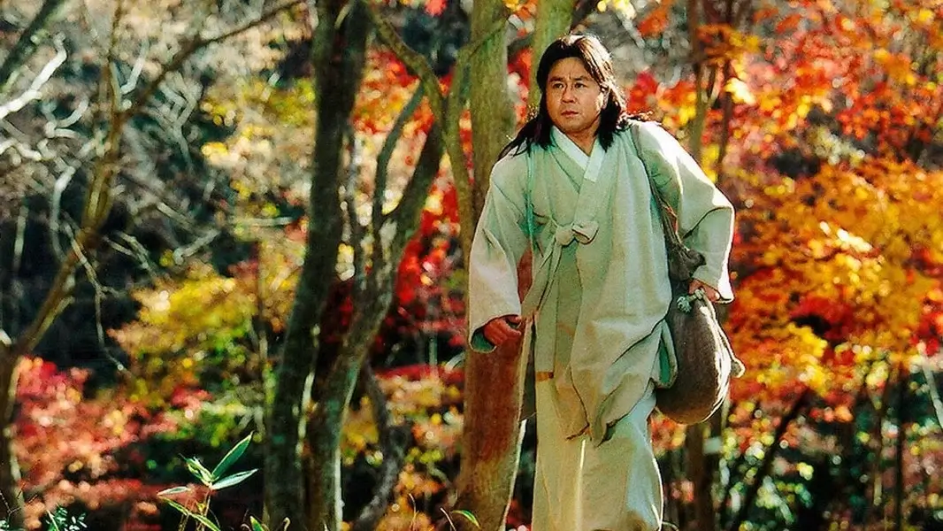 붉게 물든 가을 숲길을 따라 흰 한복 차림의 남성이 보따리를 메고 걸어가는 영화 취화선의 장면.