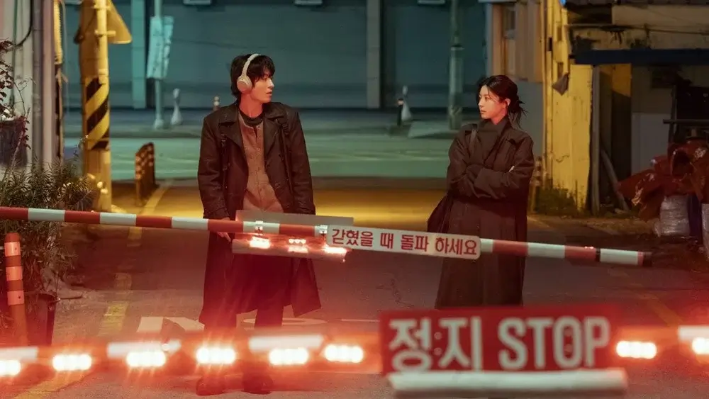 드라마 모두가 자신의 무가치함과 싸우고있다 스틸 이미지로, 남자 주인공 캐릭터를 연기하는 구교환배우와 여자 주인공 캐릭터를 연기하는 고윤정배우가 차단기 앞 골목에서 서로를 바라보며 미묘한 감정선을 형성하는 장면.