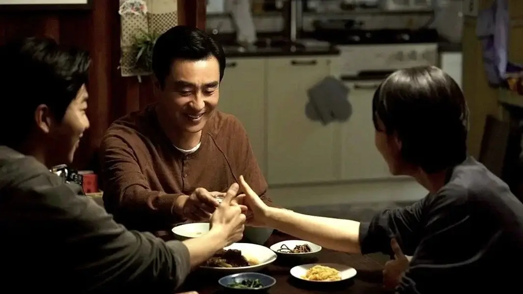 서울 자가에 대기업 다니는 김 부장 이야기 영화 스틸 이미지로, 가족이 식탁에 둘러앉아 손을 맞대는 모습이 일상적인 저녁 식사 속 따뜻한 가족 관계와 소박한 행복을 담아낸 장면.