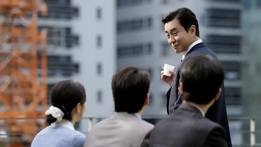 서울 자가에 대기업 다니는 김 부장 이야기 영화 스틸 이미지로, 정장을 입은 남성이 옥상에서 동료들에게 음료를 들고 이야기하는 모습이 회사 생활 속 여유와 미묘한 관계의 긴장을 함께 보여주는 장면.